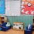 A classroom reading corner with two blue chairs, a low bookshelf, and a daily schedule bulletin board.