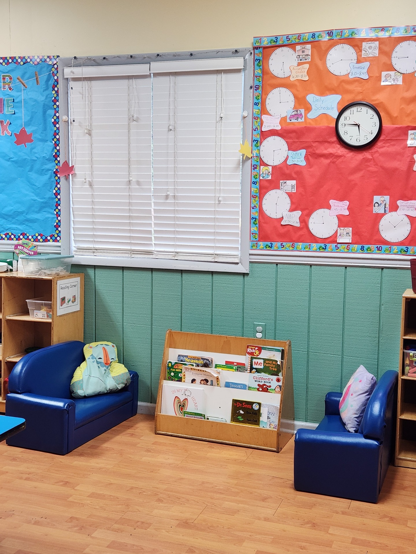 A classroom reading corner with two blue chairs, a low bookshelf, and a daily schedule bulletin board.