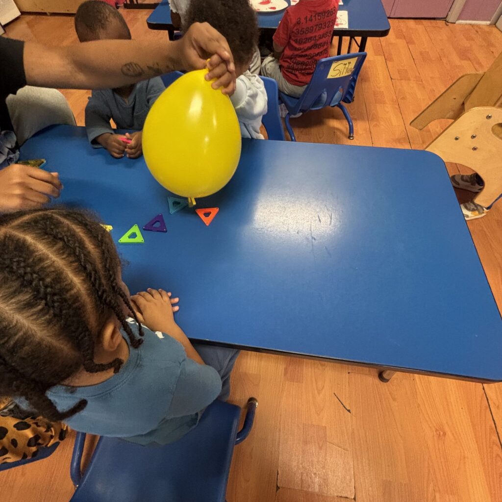 arlc-classroom-children-exploring-yellow-balloon-at-blue-table-02 Children exploring shapes and a yellow balloon with their teacher
